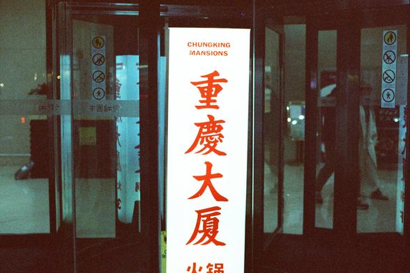 CineStill 800T (FF)

English Alt Text
A nighttime street-level view of the entrance to Chungking Mansions. A tall vertical sign glows with red Chinese characters above the English name “CHUNGKING MANSIONS.” The glass doors below display several prohibition icons, such as no smoking, no pets, and no skateboarding. The lighting is dim except for the bright signage, giving the scene a moody, urban atmosphere.
Chinese Alt Text
夜间的重庆大厦入口。高高的竖直招牌以红色中文字符发光，上方有英文“CHUNGKING MANSIONS”。下方的玻璃门贴有多个禁止标志，如禁止吸…