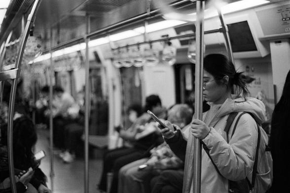 Rollei RPX 400 (FF)

English Alt Text
A black‑and‑white photo taken inside a subway car. Several passengers are seated or standing, all absorbed in their smartphones. In the foreground, a person wearing a light jacket and a backpack stands while holding a vertical pole with one hand and using a phone with the other. Overhead handles, metal poles, and a digital display screen are visible. The scene captures a typical urban commute and the quiet, absorbed atmosphere of riders focused on their dev…