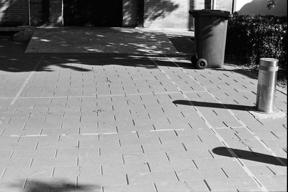 Rollei RPX 400 (FF)

English Alt Text
A black‑and‑white photograph of a paved walkway made of rectangular tiles. A cylindrical metal bollard stands in the foreground, casting a long shadow across the tiles. A wheeled trash bin sits near the edge of the walkway. In the background is a brick building with a dark doorway. Tree branches cast patterned shadows over the ground, creating a geometric, high‑contrast composition.
中文替代文字
一张黑白照片，展示由矩形砖块铺成的人行道。前景有一个圆柱形金属路桩，影子在地面上拉得很长。旁边放着一个带轮垃圾桶。背景是一栋带砖墙和暗色…