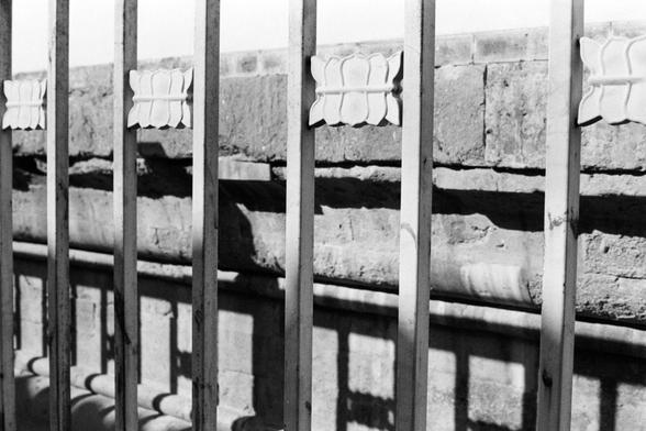 Rollei RPX 400 (FF)

English Alt Text
A black‑and‑white close‑up of a metal fence with vertical bars and small decorative leaf‑shaped ornaments attached to them. Strong sunlight casts sharp, repeating shadows of the fence onto a stone wall behind it. The wall is made of large rectangular blocks, and the contrast between the smooth metal and rough stone creates a rhythmic, geometric pattern.
Chinese Alt Text
一张黑白特写照片，画面中是带有竖条和叶片状装饰的金属栅栏。强烈的阳光将栅栏的影子清晰地投射在后方的石墙上。石墙由大块矩形石块组成，金属的光滑质感与石头的粗糙纹理形成鲜明对比，呈…