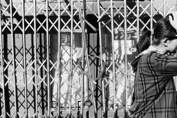 Rollei RPX 400 (FF)

English Alt Text
A black‑and‑white image of a woman standing beside a metal gate with a crisscross pattern. The gate casts dramatic, angular shadows on the wall behind it. The woman stands on the right side, facing away, with one hand resting on her head. She wears a plaid jacket, and behind the gate are posters with numbers and text. The center of the gate is bent inward, forming a heart‑like shape.
Chinese Alt Text
一张黑白照片，一名女子站在带有交叉图案的金属门旁。金属门在后方墙面上投下强烈而锐利的阴影。女子位于画面右侧，背对镜…