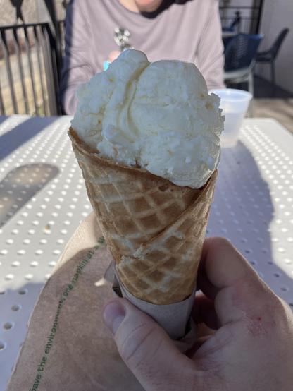 A hand holds a cone filled with a large scoop of creamy white ice cream. 
