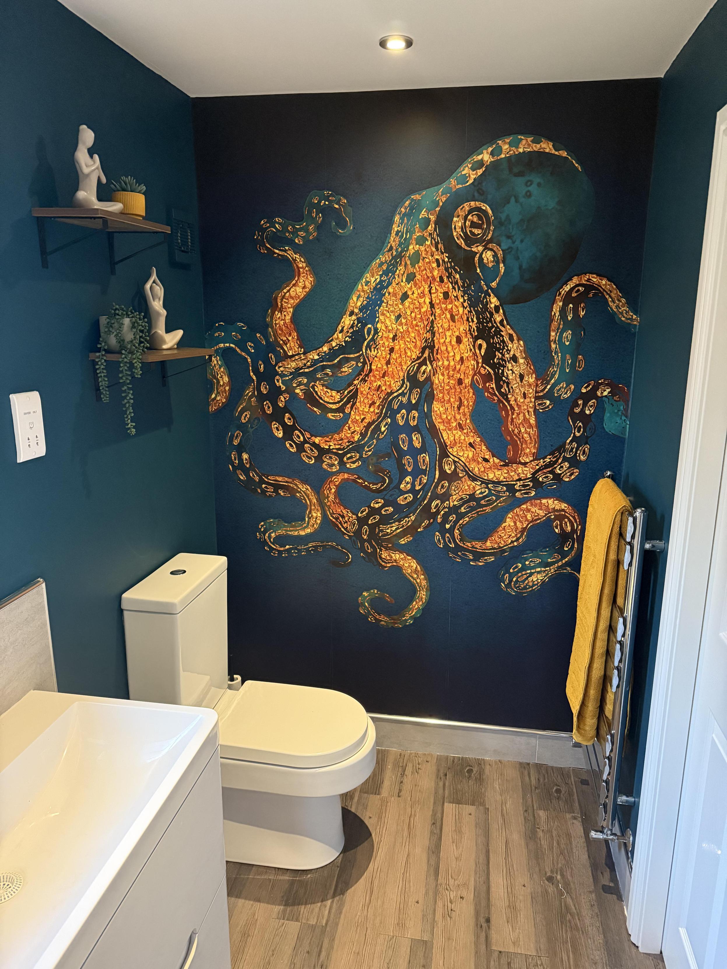 From reddit/r/HomeDecorating, somebody has put a large gold and blue octopus decal on their blue bathroom wall right next to the toilet. On shelves over the toilets you see some fake plants and two ceramic figurines of nude women meditating.