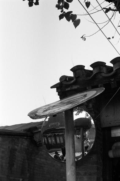 Rollei RPX 400 (FF)

Image 1 — Alt Text (English)
A black‑and‑white street photograph featuring a convex traffic mirror mounted on a pole. Above the mirror is a worn traffic sign. Reflected in the mirror is a traditional building with ornate roof details, several pedestrians, and another pole. Tree branches and part of a tiled roof frame the upper portion of the image. The scene blends old architectural elements with modern street infrastructure.
Image 1 — Alt Text (Chinese)
一张黑白街景照片，画面中心是一面安装在…