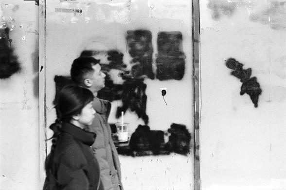 Rollei RPX 400 (FF)

Image 3 — Alt Text (English)
A black‑and‑white street scene showing two people walking past a wall covered in dark spray‑paint marks and smudges. One person holds a drink with a straw. The wall behind them is rough and weathered, with a small hole and a wire protruding from it. The overall atmosphere is gritty and urban, capturing a candid moment of daily life.
Image 3 — Alt Text (Chinese)
一张黑白街景照片，两个人从一面布满喷漆痕迹和污渍的墙前走过，其中一人手里拿着带吸管的饮料。墙面粗糙破旧，有一个小洞和一根伸出的电线。整体氛围带有都市的粗粝感，呈现日常生活…