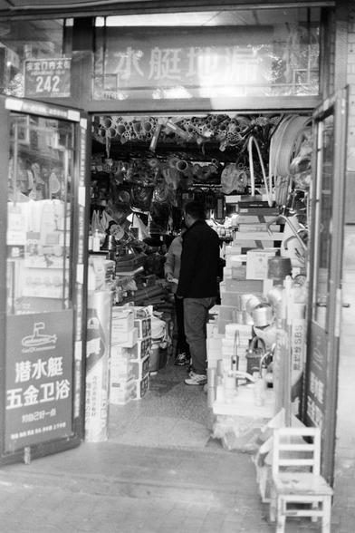 Rollei RPX 400 (FF)

Image 4 — Alt Text (English)
A black‑and‑white photograph of a crowded hardware store entrance filled with plumbing supplies. Pipes, hoses, boxes, and various fittings are stacked densely. Two people inside appear to be talking or making a transaction. Chinese signage above the entrance advertises plumbing and hardware products, and the address “Andingmen Inner Street No. 242” is visible. The scene captures the busy, cluttered character of a small urban shop.
Image 4 — Alt …
