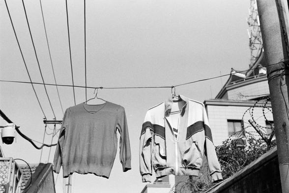 Rollei RPX 400 (FF)

English alt text
A black‑and‑white outdoor scene showing two garments hanging from a clothesline on wire hangers. On the left is a long‑sleeved sweater; on the right, a sporty zip‑up jacket with stripes. In the background are utility poles, wires, a security camera, and a building with windows and rooftop structures, blending domestic life with urban infrastructure.
中文替代文字
一张黑白户外照片，两件衣服挂在晾衣绳上。左边是长袖毛衣，右边是带条纹的运动外套。背景中可见电线杆、电线、监控摄像头以及一栋带窗户和屋顶结构的建筑，呈现家庭生活与城市环境的交织。