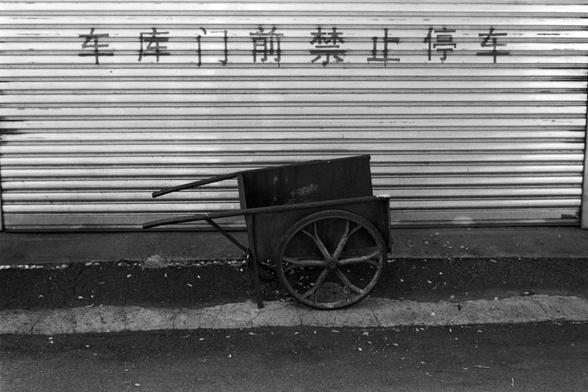 Rollei RPX 400 (FF)

English alt text
A black‑and‑white photo of an old metal handcart with two large spoked wheels and long handles. It sits directly in front of a corrugated metal garage door. The cart looks worn, with rust and scratches. Above it, bold Chinese characters painted on the door state that parking is forbidden. The ground is cracked and slightly littered, giving the scene a gritty, urban feel.
中文替代文字
一张黑白照片，画面中一辆老旧的金属手推车停在卷帘式车库门前。手推车有两个大轮子和长把手，表面布满磨损和锈迹。车库门上方写着“车库门前禁止停车”。地面有裂缝和碎屑…
