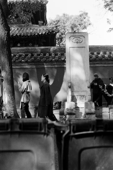 Rollei RPX 400 (FF)

English Alt Text:
A black-and-white street photograph shows people gathered near a stone monument or stele in front of a traditional Chinese wall with ornate roof tiles. Some individuals are walking, while one person appears to be taking a photograph and another bends down, possibly picking something up. The background reveals a pagoda-style building partially hidden by trees. Shadows fall across the wall and ground, creating texture. In the foreground, large plastic bins s…