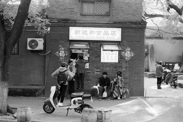 Rollei RPX 400 (FF)

English Alt Text:
A black-and-white photograph depicts a small food shop with a sign reading “新驰和食品店,” meaning “Xin Chi He Food Store.” Below, another sign lists items such as drinks and snacks with prices. Several people stand at the window ordering food, while others sit on the ledge outside. A scooter is parked in the foreground, and an air conditioning unit is mounted on the wall. One person wears traditional Chinese attire, adding cultural richness. The scene blends ev…