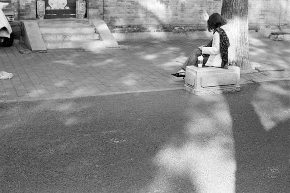 Rollei RPX 400 (FF)
English Alt Text:
A black-and-white photograph of a quiet urban street. A person sits alone on a stone bench near a tree, with a coffee cup placed beside them. They are focused on a mobile device, suggesting a moment of solitude or reflection. The background shows a brick wall and stone steps leading to a decorative doorway. Shadows from trees fall across the ground, creating patterns on the pavement. The atmosphere feels calm, contemplative, and still, capturing everyday li…