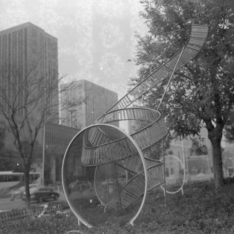 Ilford HP5 Plus 400 (6x6)

Alt Text (English)
A black‑and‑white photo of a large abstract metal sculpture in a city setting. The sculpture is made of circular and spiral shapes resembling a twisted ribbon, supported by metal frames. It sits among trees and bushes. In the background are tall office buildings and a street with moving vehicles, including a bus.

Alt Text (Chinese)
一张黑白照片，主体是一座大型抽象金属雕塑，由圆形和螺旋形结构组成，像扭曲的金属带，由支架支撑。雕塑周围有树木和灌木。背景是高楼和有车辆经过的街道，包括一辆公交车。