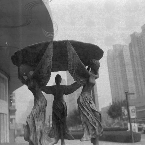 Ilford HP5 Plus 400 (6x6)

Alt Text (English)
A black‑and‑white photo of an outdoor sculpture featuring three winged female figures. They stand close together, lifting a large circular disc above their heads. Their wings and flowing garments create a sense of motion. Behind them are tall modern buildings and a curved architectural structure. The image has a vintage, film‑like grain.

Alt Text (Chinese)
一张黑白照片，画面中有一座户外雕塑，由三位长着翅膀的女性形象组成。她们紧靠在一起，高举一个大型圆盘。人物的翅膀和飘动的衣饰呈现出动态感。背景是高楼与弧形的现代建筑。整体带有胶片质感。