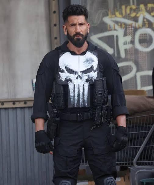 A muscular, bearded Frank Caslte stands facing the camera in a black tactical outfit with gloves and knee pads, wearing a vest marked by a large white skull emblem. He’s in a gritty, industrial-looking area with graffiti and a metal shopping cart behind him, giving the scene a tough, urban, action-movie vibe.