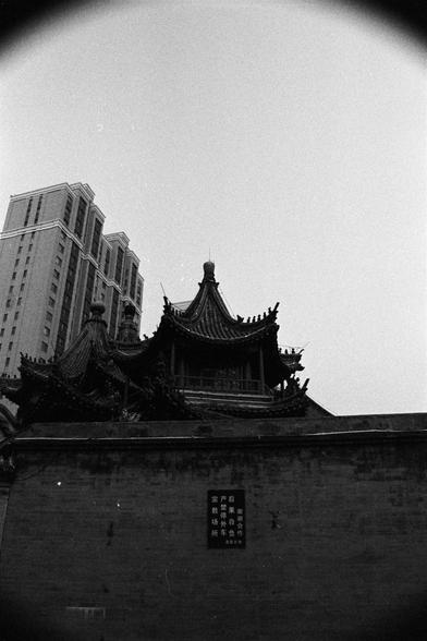 Rollei RPX 400 (FF)

English Alt Text:
A black-and-white photograph showing a traditional Chinese temple in the foreground. Its ornate rooftops curve upward with decorative details, representing religious and cultural heritage. Behind the temple, a modern high-rise building towers, symbolizing urban growth. A sign on the temple wall in Chinese warns: “Consequences are at your own risk. External vehicles are strictly prohibited. Religious site. Thank you for your cooperation.” The image captures…