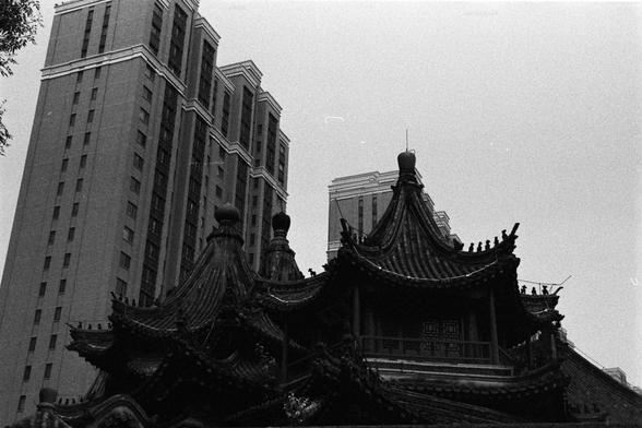 Rollei RPX 400 (FF)

English Alt Text:
A photograph showing the contrast between old and new architecture. In the foreground, a traditional East Asian building with ornate, curved rooftops and intricate wooden carvings stands proudly. The roof ridges are decorated with small figurines and finials, emphasizing classical Chinese design. Behind this historic structure, tall modern skyscrapers rise with clean lines, glass windows, and geometric patterns. The juxtaposition highlights the coexistence…