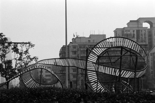 Rollei RPX 400 (FF)

English Alt Text:
A black-and-white photograph of a large abstract metal sculpture in a public space. The sculpture is made of curved and twisted metal slats arranged in a looping, ribbon-like form, resembling a Möbius strip or roller coaster track. Supported by a sturdy frame, the structure conveys motion and complexity. In the background, residential buildings and trees frame the scene, contrasting the organic curves of the artwork with the rigid geometry of urban archite…