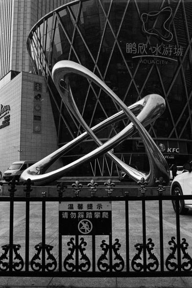Rollei RPX 400 (FF)

English Alt Text:
A large, futuristic sculpture stands in front of a commercial building named Aqua City. The sculpture consists of two intertwined metallic loops with a reflective surface, resembling a dynamic ribbon. Behind it, the building’s glass facade features a diamond-shaped pattern and displays logos of international brands such as KFC and Prada. A decorative fence in the foreground carries a sign in Chinese, politely reminding visitors not to step on or climb. The…