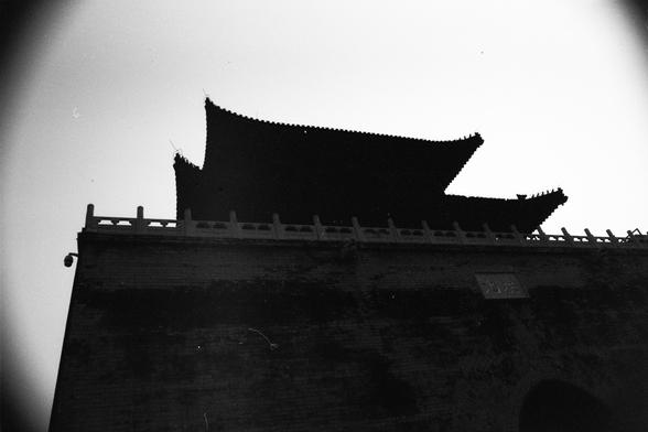 ERA 100 (FF)

Image 4 – English Alt Text
A black‑and‑white photograph of a traditional Chinese gate or tower with a multi‑tiered roof and upturned eaves. The structure stands on a brick base with a railing along the top. The building is silhouetted against a bright sky, highlighting the roof’s layered curves. A sign with Chinese characters is visible on the brick wall.
Image 4 – 中文替代文字
一张黑白照片，呈现一座传统中国式城门或楼阁，屋顶多层叠起并带有飞檐。建筑立于砖石基座上，顶部有栏杆。建筑在明亮天空前形成剪影，屋檐的层次清晰可见。砖墙上可见一块写有中文的牌匾。