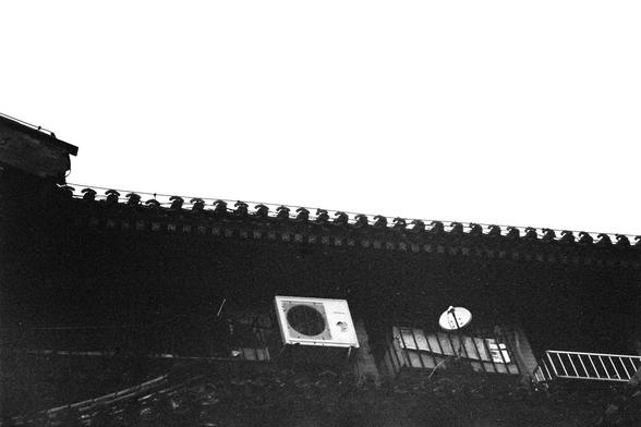 ERA 100 (FF)

Image 1 – English Alt Text
A black‑and‑white photo of the upper part of a traditional East Asian building. The roof has ornate tiles and decorative ridge details. Below the roofline, a modern air‑conditioning unit and a satellite dish are mounted on the wall, creating a contrast between old architecture and contemporary technology. The lighting is soft, and the textures of the tiles and wall are clearly visible.
Image 1 – 中文替代文字
一张黑白照片，呈现一座传统东亚建筑的上半部分。屋顶铺有精致的瓦片和装饰性的屋脊。屋檐下安装着现代的空调机…