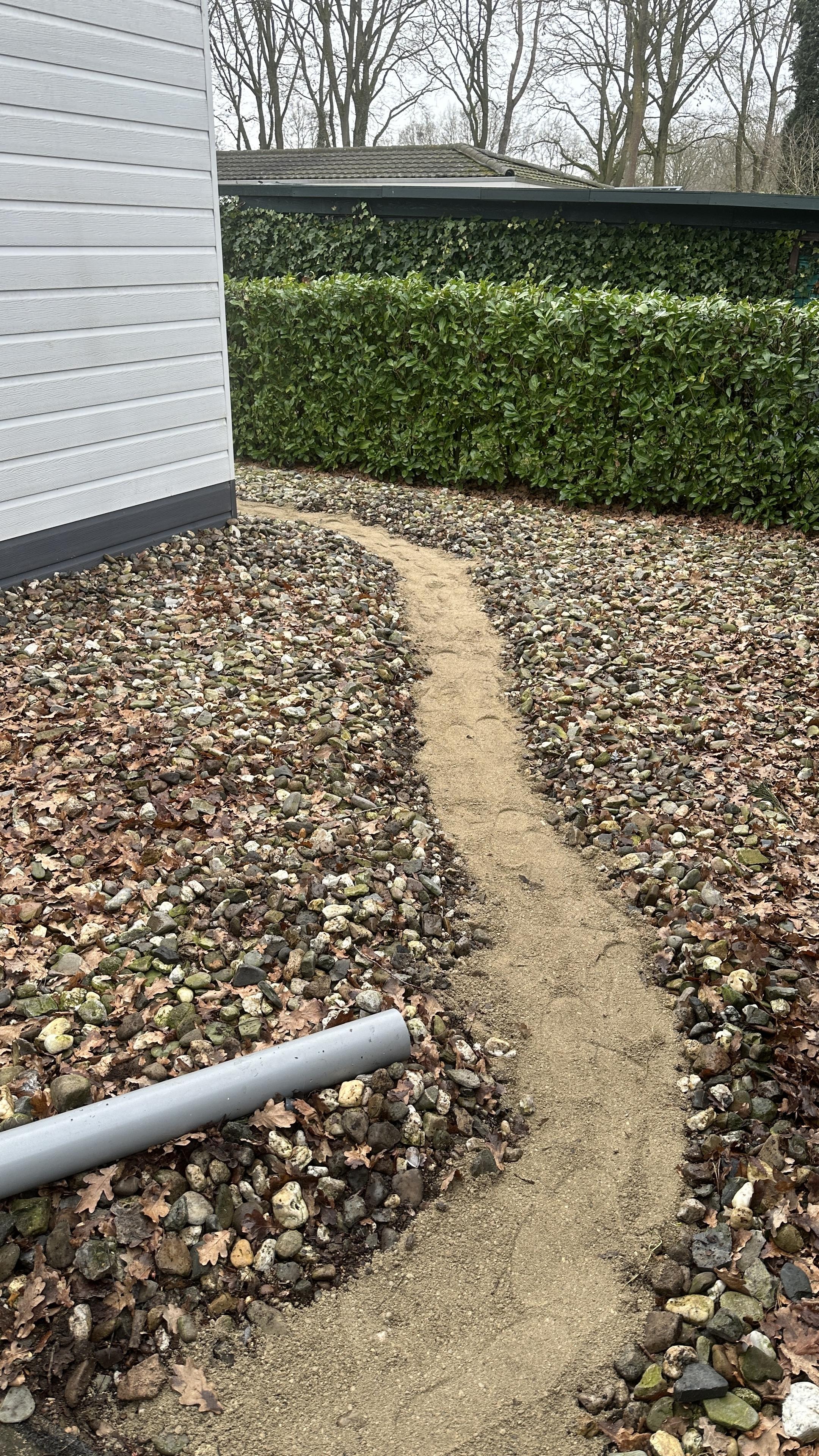 A narrow dirt path winds through a gravel-covered yard, leading beside a white house with green hedges and leafless trees in the background.