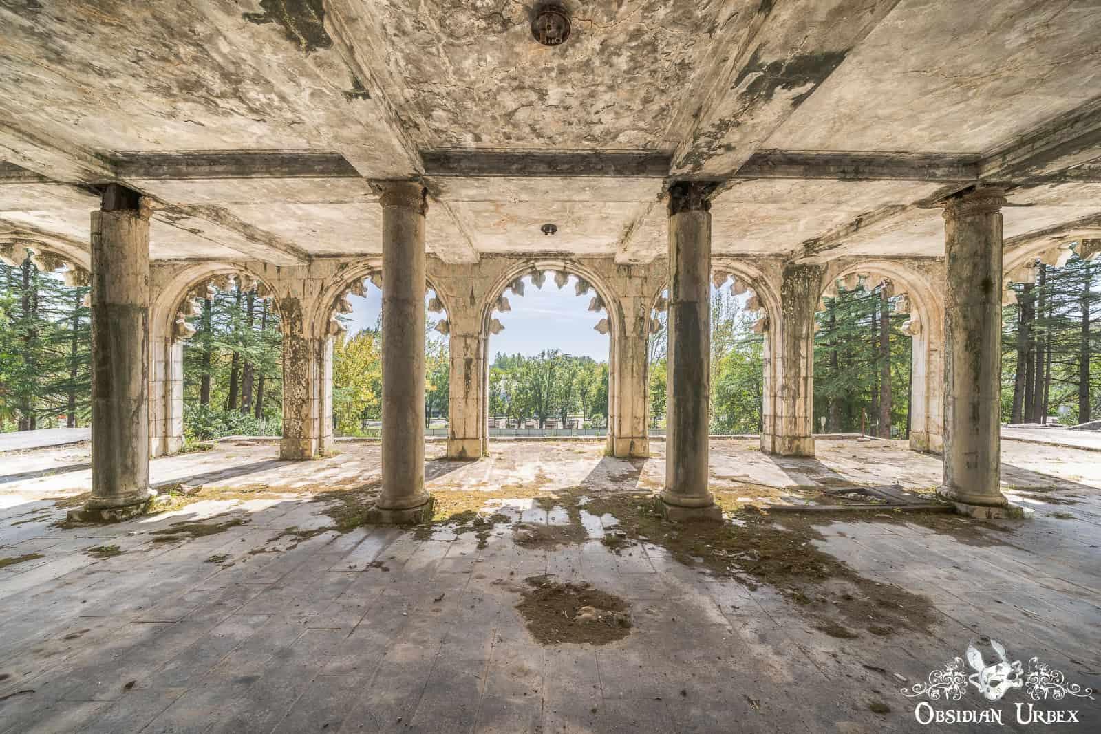 An abandoned, ornate concrete hall features multiple columns and scalloped arches opening to trees.