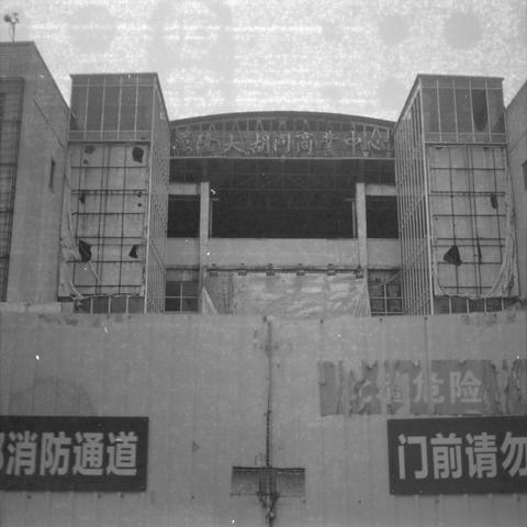 Ilford HP5 Plus 400 (6x6)

English alt text:
A black‑and‑white photo of a damaged multi‑story commercial building. The central section is hollow and exposed, with missing walls and visible structural beams. The two vertical side sections have torn exterior panels hanging loosely. At the top is a sign in Chinese characters indicating the name of the commercial center. At street level, additional signs appear, including one marking a fire passage. The overall scene shows a deteriorated, abandoned…