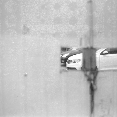 Ilford HP5 Plus 400 (6x6)

English alt text:
A grainy black‑and‑white image showing several parked cars in a lot, viewed through a translucent or frosted foreground surface. A vertical object partially blocks the view. Only a small clear section reveals a sharply focused white car, creating a layered contrast between blurred foreground and crisp background.
中文替代文字：
一张黑白颗粒感照片，通过半透明或磨砂状的前景表面看到停车场中的多辆汽车。前景有一条垂直物体遮挡部分视线。只有一小块清晰区域能看到一辆白色汽车，形成模糊前景与清晰背景的对比层次。
