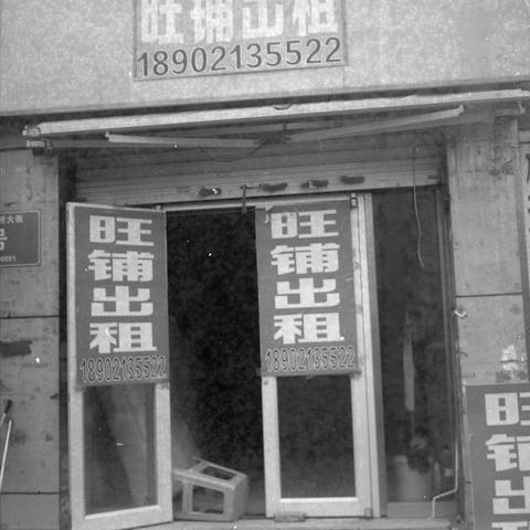 Ilford HP5 Plus 400 (6x6)

English alt text:
A black‑and‑white image of an empty storefront with multiple signs reading “Prime Shop for Rent” in Chinese, along with a phone number. The signs appear above the entrance and on the open doors. Inside the vacant space, a few items such as a crate and a mop are visible.
中文替代文字：
一张黑白照片，画面为一间空置商铺，门上方和敞开的门板上挂着多张写有“旺铺出租”和电话号码的招租告示。店内空荡，可见塑料筐和拖把等零散物品