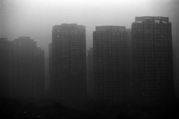 ERA 100 (FF)
English Alt Text:
A black and white photograph shows a cluster of high-rise apartment buildings. The towers are closely packed, but their details are blurred by thick smog or fog. The atmosphere is dark and heavy, with little visibility. The lack of clarity emphasizes urban density and environmental challenges, evoking a somber mood of pollution or obscured city life.

中文替代文字：
这是一张黑白照片，画面是一组高层住宅楼。建筑紧密排列，但因浓雾或雾霾而模糊不清。整体氛围阴沉、能见度低。画面突出了城市的密集感和环境问题，传递出一种因污染或迷雾而显得沉重的城市氛围。