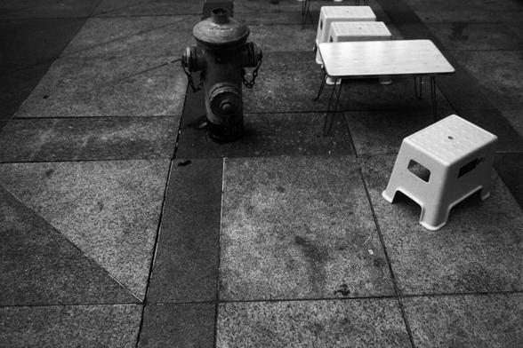 ERA 100 (FF)

English Alt Text:
A black and white photo shows a paved sidewalk or plaza with geometric tiles. In the center stands a fire hydrant, its side nozzles secured with short chains. To the right, several small plastic stools and tables are arranged in a line, suggesting a casual outdoor seating area. The contrast between the utilitarian hydrant and the makeshift furniture creates a structured yet informal urban scene.

中文替代文字：
这是一张黑白照片，画面是一片铺设方形和三角形地砖的人行道或广场。中央有一个消防栓，两侧的接口用短链条固定。右侧摆放着几…