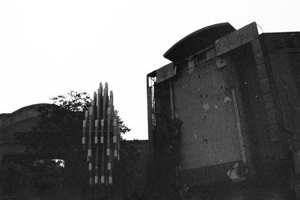 ERA 100 (FF)

English Alt Text:
A black and white photograph depicts a large, weathered building with a tall vertical facade. The surfaces show peeling and broken details, suggesting age or neglect. To the left, a striking vertical sculpture made of clustered cylindrical forms rises, resembling organ pipes or a modernist installation. Trees are faintly visible behind, contrasting with the industrial decay. The scene conveys urban history and architectural tension.

中文替代文字：
这是一张黑白照片，画面是一座高大的旧建筑，…