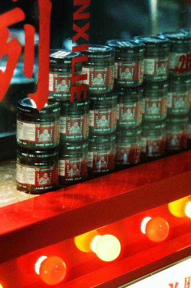CineStill 800T (FF)

English:
This image shows a close-up of a store shelf displaying stacks of round jars. The jars are black with red and white labels featuring an image of a person’s face and Chinese text. The jars are neatly arranged in rows and columns, and the word "OPEN" is visible on the lids. The background includes a red and white sign with partial Chinese characters, and the lighting is warm, creating a cozy atmosphere.

Chinese:
这张图片展示了商店货架上整齐摆放的圆形罐子。罐子为黑色，贴有红白相间的标签，标签上印有一张人物脸部图片和中文…