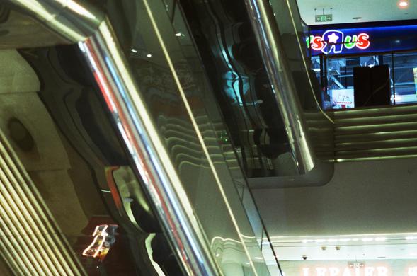 CineStill 800T (FF)

English:
This image captures an escalator with a reflective metal railing. The escalator is in a shopping mall, and the reflection on the railing shows the mall's interior, including a Toys"R"Us store sign in the background. The lighting is bright, and the mall appears spacious and modern.

Chinese:
这张图片展示了购物中心内的自动扶梯，扶梯的金属扶手反射出商场内部的景象。扶手的反射中可以看到远处的玩具反斗城（Toys"R"Us）店招。商场内灯光明亮，空间宽敞，现代感十足。