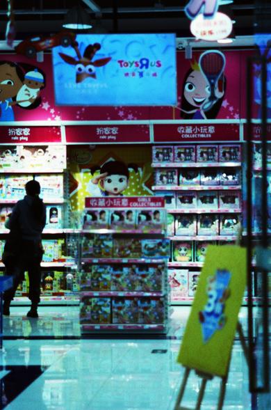 CineStill 800T (FF)

English:
This image captures the interior of a toy store, specifically the Toys"R"Us brand. The scene shows shelves filled with various toys and collectibles, including dolls and action figures. The shelves are well-lit, and colorful packaging is visible. A person is standing in front of the shelves, browsing the items. Above the shelves, there are signs in both English and Chinese, indicating different sections like "role play" and "collectibles."

Chinese:
这张图片展示了玩具反斗城（To…