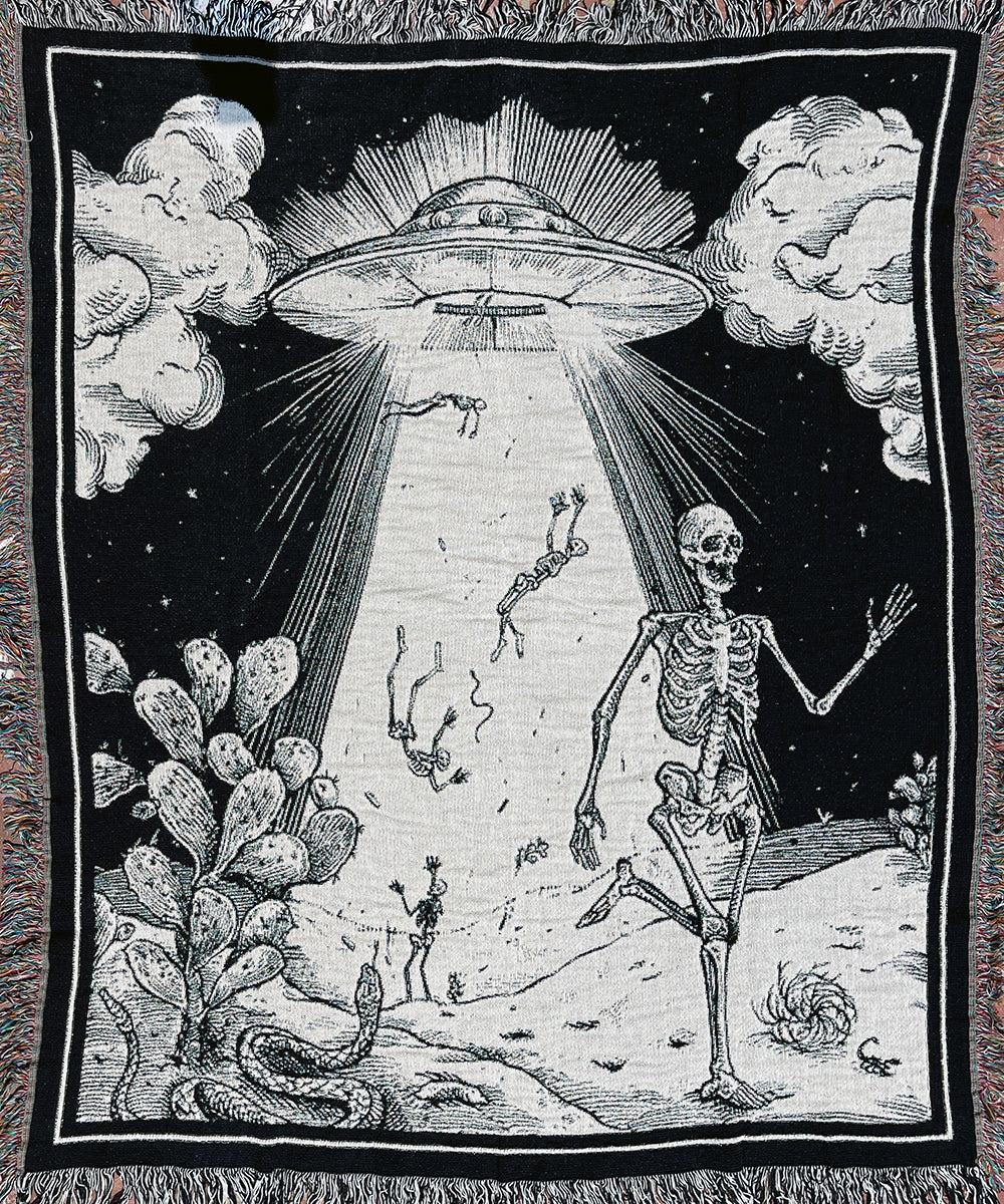 A black and white woven blanket showing a large UFO beaming up skeletons from a desert scene with cactus and a snake.