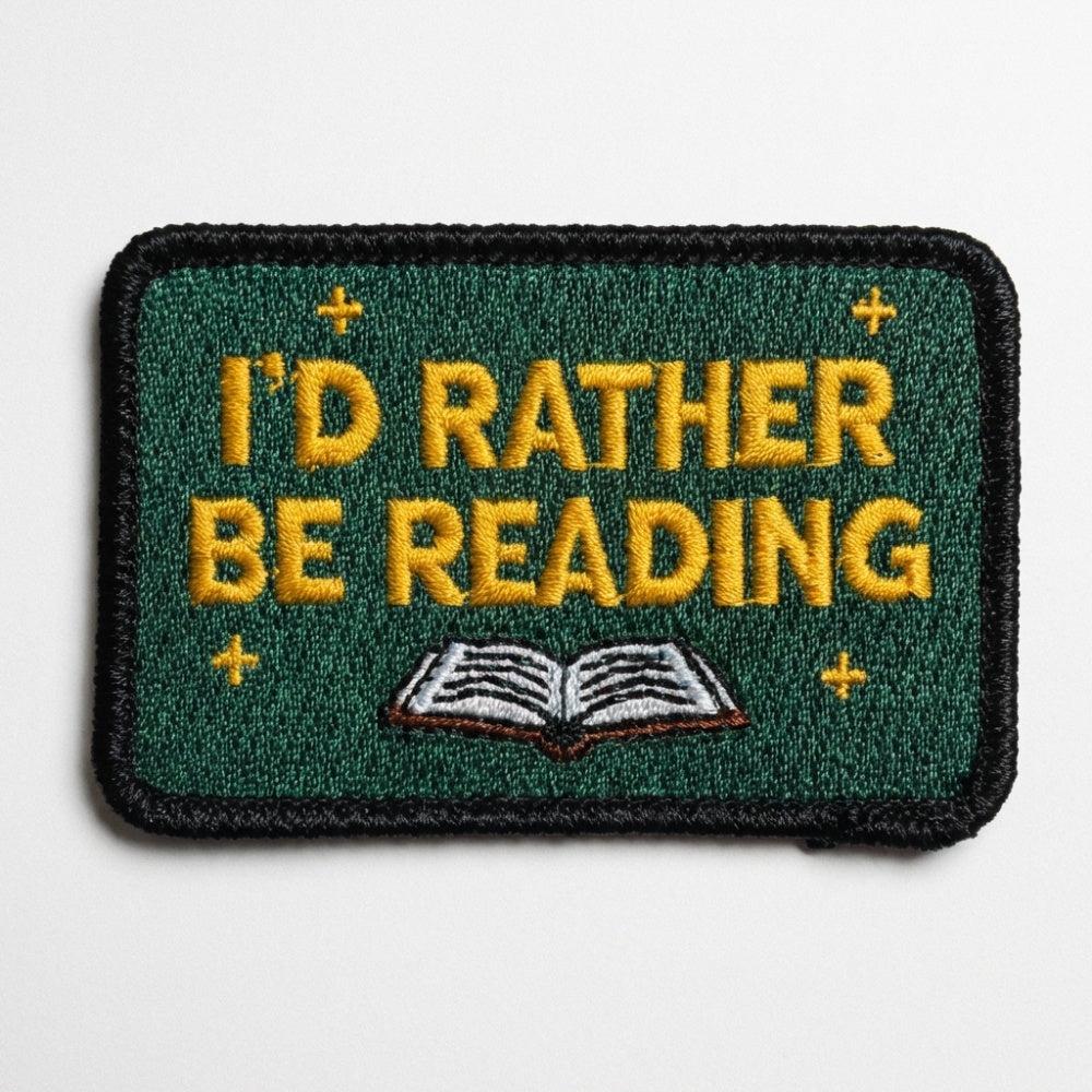 A rectangular green embroidered patch with yellow text reading "I'D RATHER BE READING" above an open book illustration.