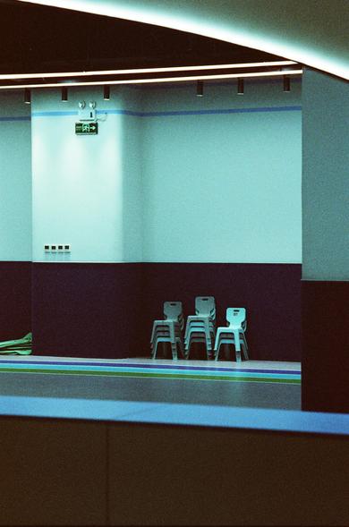 CineStill 800T (FF)

English Alt Text
An indoor space with a two‑tone blue wall, light blue on top and dark blue on the bottom. Five stacked plastic chairs sit in the corner beneath an emergency exit sign with Chinese characters and a left‑pointing arrow. The ceiling has integrated lighting strips, and the floor features colorful stripes in blue, green, and purple. Electrical outlets and coiled green cables rest along the left wall, giving the room a clean but utilitarian feel.
中文替代文字
一个室内空间，墙面…