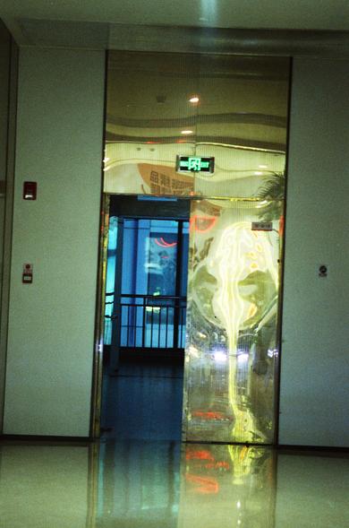CineStill 800T (FF)

English Alt Text
A hallway leading to a doorway marked by a glowing green exit sign with Chinese characters and the word “EXIT.” To the right of the doorway is a reflective panel showing distorted neon‑like colors in yellow, red, and blue. Beyond the doorway, a room with blue lighting and metal railings is visible. The polished floor reflects the lights, and a fire alarm and safety sign are mounted on the left wall.
中文替代文字
一条走廊通向一扇门，门上方有绿色发光的出口标志，包含中文和“EXIT”字样。门右侧的反光面呈现扭曲的黄…