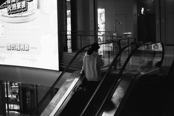 ERA 100 (FF)

English alt text:
A black‑and‑white mall scene showing a person in a hoodie riding an escalator while looking at their phone. To the left is a large, glowing advertisement with Chinese text. The surroundings include glass railings, store entrances, and overhead lighting. The bright ad contrasts sharply with the darker escalator area.

中文替代文字：
一张黑白商场照片，一位穿连帽衫的人在自动扶梯上看手机。左侧有一块发光的中文广告牌。周围有玻璃栏杆、店铺入口和顶部灯光。明亮的广告与较暗的扶梯区域形成强烈对比。