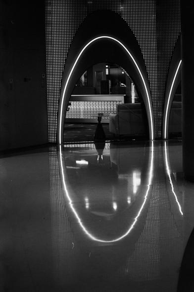 ERA 100 (FF)

English alt text:
A black‑and‑white interior scene featuring a large illuminated arch with tiny lights or perforations along its surface. The polished floor reflects the arch, forming an eye‑shaped symmetry. Inside the arch is a seating area with a couch and small table, and behind it is a counter with similar lighting. The overall style feels sleek and futuristic.

中文替代文字：
一张黑白室内照片，主体是一个带灯光的大型拱形结构，表面布满小灯点或孔洞。光亮地面映出拱形，形成眼形对称。拱内有沙发和小桌子，后方是带灯光的柜台。整体风格现代且富有未来感。