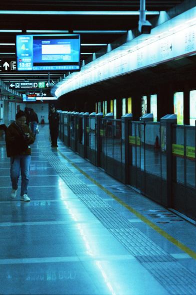 CineStill 800T (FF)

English Alt Text
A modern subway platform with glass safety doors separating passengers from the tracks. Overhead signs in Chinese and English show directions, including “Exit D,” “To Xianghu,” and “To Longxiangqiao.” A digital clock reads 13:57. The floor is polished and reflects blue lighting, with tactile paving guiding along the platform edge. A few people stand or walk: one person looks at a phone while walking, and another in a uniform stands farther down the platform…