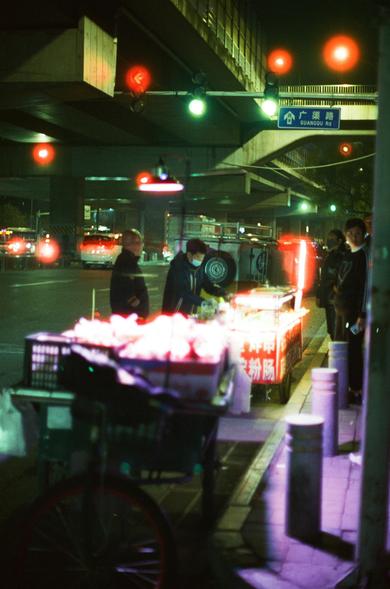 CineStill 800T (FF)

English Alt Text
A nighttime urban scene beneath an overpass, illuminated by colorful lights from traffic signals and vendor carts. Several street food stalls are set up, one glowing brightly with white and orange light. A few people stand nearby, including someone wearing a face mask. A blue bilingual street sign reading “GUANGOU Rd” hangs overhead. Light flares, reflections, and shadows create a vivid, cinematic atmosphere.
中文替代文字
夜间的城市街景位于高架桥下，由交通灯和小吃摊的彩色灯光照亮。多辆小推车式的街头摊位…