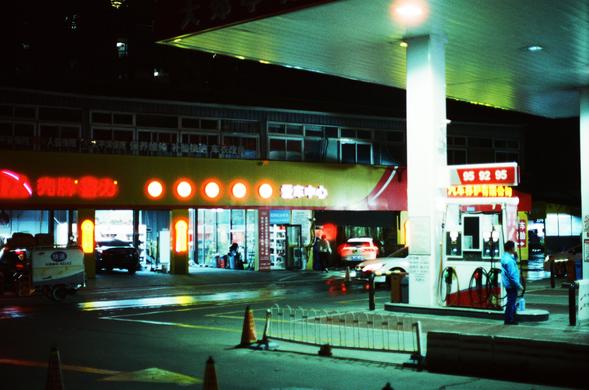 CineStill 800T (FF)

English Alt Text
A brightly lit gas station at night with a wide canopy covering several fuel pumps. A person stands beside one of the pumps, and the wet ground reflects the strong white and yellow lights. Behind the pumps is a building with illuminated Chinese signage, likely a service or repair center. One vehicle is parked outside while another is being serviced inside the open garage. Traffic cones and barriers guide cars through the area. The overall scene feels quiet,…