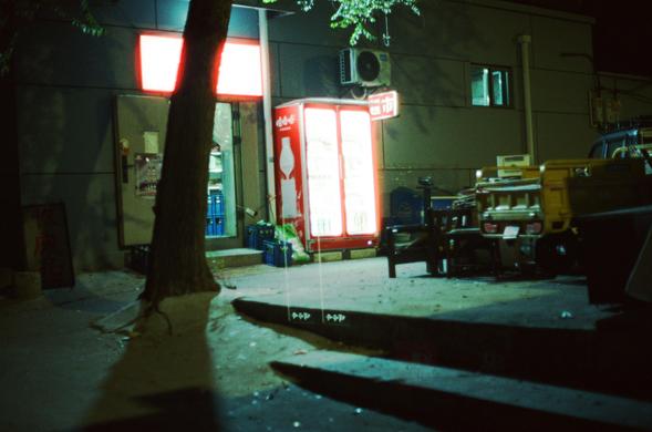 CineStill 800T (FF)

English Alt Text
A small convenience store at night with a bright red-and-white sign above the entrance. A red Coca‑Cola refrigerator stands outside, glowing with interior light and filled with visible bottles. Crates and green glass bottles sit near the doorway. A tree trunk partially blocks the view in the foreground. To the right, a yellow utility vehicle and several benches or tables are dimly lit. The surrounding street is dark except for the store’s lights, giving the…