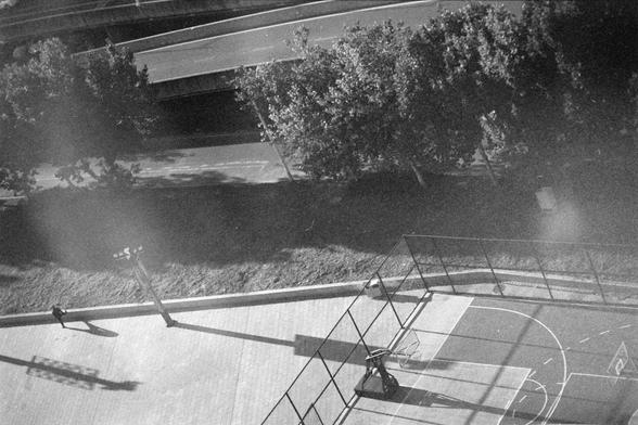 Ilford HP5 Plus 400 (FF)

English alt text:
An overhead black‑and‑white view of an outdoor basketball court next to a curved road. The court has clear boundary lines and two hoops. A single person walks near the court, casting a long shadow due to low‑angle sunlight. Tall light poles also cast long shadows across the ground. Trees surround part of the area, adding texture to the scene.
中文替代文字：
一幅从上方拍摄的黑白画面，展示一座靠近弯曲道路的户外篮球场。球场线条清晰，两端各有一个篮框。一个人走在球场旁，因阳光角度低而投下长长的影子。高大的灯柱也在地面上形成长影。周围部分区域被树木环绕。
