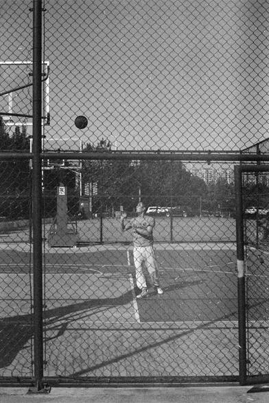 Ilford HP5 Plus 400 (FF)

English Alt Text
A black‑and‑white photo taken through a chain‑link fence shows a person on an outdoor basketball court. The person stands with arms raised, looking up toward a basketball suspended in mid‑air. Two basketball hoops, parked cars, trees, and buildings appear in the background. The fence in the foreground creates a layered perspective.
中文替代文字
一张黑白照片从铁丝网外拍摄，画面中一名站在户外篮球场上的人正抬头看向空中的篮球，双臂举起。背景有两个篮球架、停放的车辆、树木和建筑物。前景的铁丝网形成了分层的视觉效果。