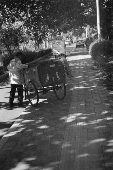 Ilford HP5 Plus 400 (FF)

English Alt Text
A black‑and‑white street scene shows a person pushing a three‑wheeled cart along a tree‑lined sidewalk. The cart has the numbers “078” on its side and carries sticks or branches. The person wears a jacket and cap. Sunlight filters through the trees, creating patches of light on the pavement. Cars and more trees line the road in the background.
中文替代文字
一张黑白街景照片中，一名穿着夹克和帽子的人正推着一辆三轮小车沿着树荫人行道前行。小车侧面写着“078”，车内装着树枝或木棍。阳光透过树叶在地面形成斑驳光影。背景可见道路上的车辆和更多树木。