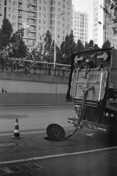 Ilford HP5 Plus 400 (FF)

English Alt Text
A black‑and‑white image shows a maintenance truck parked beside an open manhole on a city street. A large hose extends from the truck into the manhole. A traffic cone is placed nearby, and the manhole cover rests on the ground. Tall residential buildings and trees line the street in the background.
中文替代文字
一张黑白照片显示一辆维护车辆停在城市道路旁的敞开井盖旁。粗大的软管从车上延伸进入井口。附近放置了一个交通锥，井盖靠在地面上。背景是高层住宅楼和街道两侧的树木。