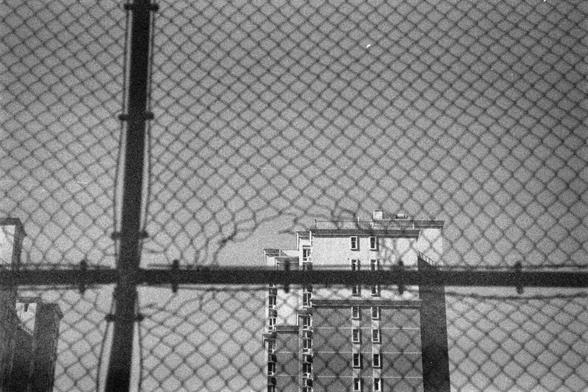Ilford HP5 Plus 400 (FF)

Image 1 – Alt Text (English)

A black‑and‑white photo showing a tall building seen through a chain‑link fence. The fence is close to the camera and has a bent or damaged opening in the center. Through this opening, a multi‑story building with many windows is visible in the background. The building appears rectangular and modern. The angle is slightly low, making the building look taller. The focus is on the fence, while the building behind it is somewhat softer in deta…